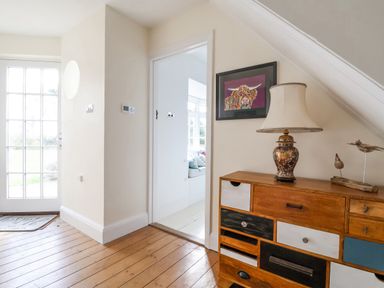 A hallway with a wooden floor a door to the outside a multicolored drawer cabinet with a lamp and bird figurines and an open door to a bright room at Halsey in Walberswick