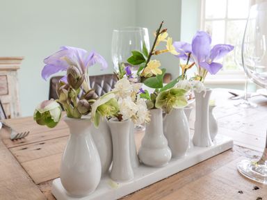 A wooden table with multiple small white vases holding various flowers and wine glasses at Halsey in Walberswick