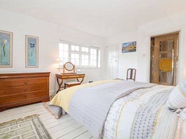 A bedroom with a bed against the right wall a wooden dresser two floral paintings a vanity table with a mirror and lamp a chair and a doorway to a bathroom at Halsey in Walberswick