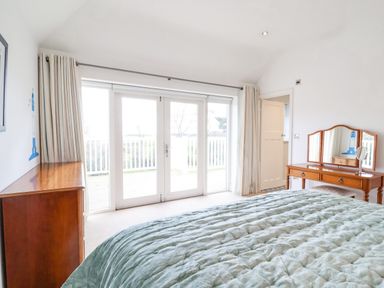 A bedroom with a bed covered in a quilted blanket wooden dresser and vanity table with mirrors near glass doors leading outside at Halsey in Walberswick