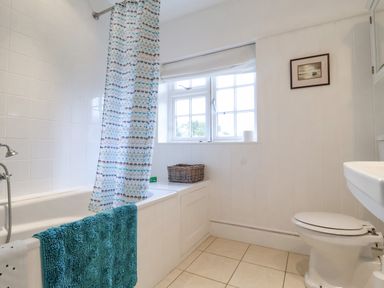 A bathroom with a bathtub and shower curtain a window with a wicker basket a toilet and a white sink at Halsey in Walberswick