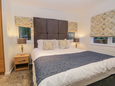A bedroom with a bed with patterned cushions and gray headboard bedside tables with lamps and windows with patterned blinds at 25 Thirlmere in Troutbeck Bridge