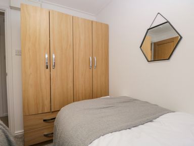 A bedroom with a wooden wardrobe and a bed with a gray blanket and a hexagonal mirror on the wall at 25 Thirlmere in Troutbeck Bridge