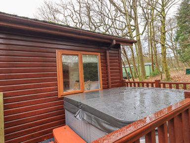 A hot tub with a cover on a wooden deck outside a log cabin with trees in the background at 25 Thirlmere in Troutbeck Bridge