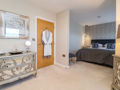 A bedroom with a bed with pillows and a decorative wallpaper behind it a door with robes hanging and a dresser with a mirror and decorative items at The Penthouse in Woodbridge