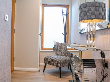 A small room with a patterned chair next to a window and a mirrored table with a lamp on top at The Penthouse in Woodbridge