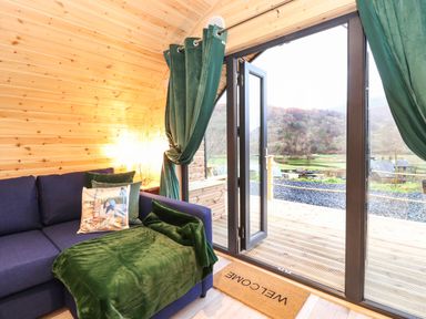 A wooden interior room with a blue sofa and green curtains next to open glass doors opening to an outdoor deck at The Shearer - Crossgate Luxury Glamping in Hartsop near Glenridding
