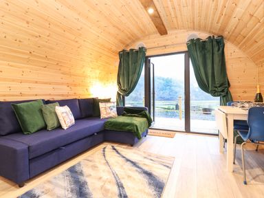 A living area with a blue sectional sofa with green cushions a rug and glass doors with green curtains at The Shearer - Crossgate Luxury Glamping in Hartsop near Glenridding