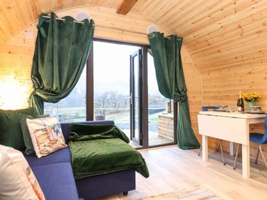 A small wooden room with a blue sofa, green curtains, a white dining table with blue chairs and glass doors leading outside at The Shearer - Crossgate Luxury Glamping Hartsop near Glenridding