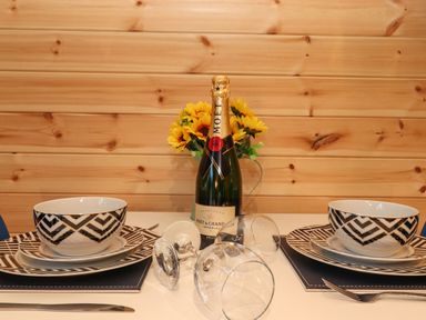 A dining table set for two with patterned bowls and plates a bottle of champagne flowers and two wine glasses at The Shearer - Crossgate Luxury Glamping Hartsop near Glenridding