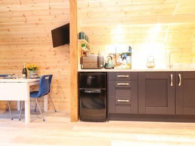 A kitchen area with a small black fridge microwave kettle and dark cabinets next to a dining table with blue chairs and dishes at The Shearer - Crossgate Luxury Glamping Hartsop near Glenridding