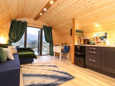 A living area with a blue sofa, green curtains, a small dining table with two chairs, and a kitchenette at The Shearer - Crossgate Luxury Glamping in Hartsop near Glenridding