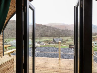 A view through open double doors onto a wooden deck with rope railing and a picnic bench overlooking fields hills and a house at The Shearer - Crossgate Luxury Glamping in Hartsop near Glenridding