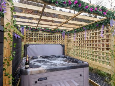 An outdoor hot tub under a wooden pergola decorated with artificial flowers and string lights at The Shearer - Crossgate Luxury Glamping in Hartsop near Glenridding