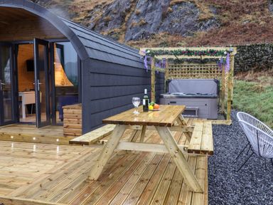 An outdoor wooden deck with a picnic table a bottle and glasses near a black curved cabin and a hot tub with a pergola at The Shearer - Crossgate Luxury Glamping Hartsop near Glenridding
