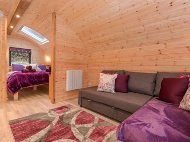 A room with wooden walls a gray sofa with pillows a patterned rug and a bed with purple bedding at Lovies Place - Crossgate Luxury Glamping in Hartsop near Glenridding