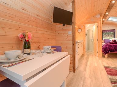 A small dining table with plates glasses and a champagne bottle in a wooden cabin with kitchen bathroom and bed visible at Lovies Place - Crossgate Luxury Glamping in Hartsop near Glenridding