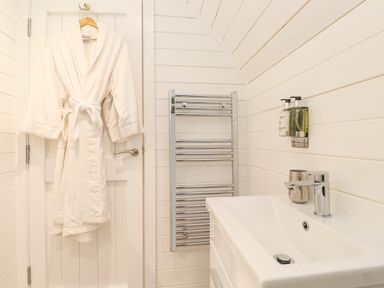 A white bathrobe hanging on a door next to a chrome towel rack and a bathroom sink with soap dispensers at Lovies Place - Crossgate Luxury Glamping in Hartsop near Glenridding