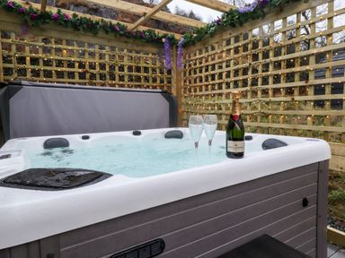 An outdoor hot tub with a bottle of champagne and two glasses on the edge surrounded by wooden lattice with string lights and floral decorations at Lovies Place - Crossgate Luxury Glamping in Hartsop near Glenridding