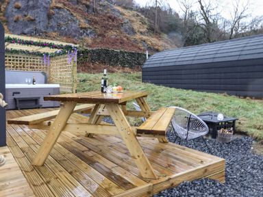 An outdoor wooden picnic table on a deck with a wine bottle and fruit beside a hot tub and fire pit at Lovies Place - Crossgate Luxury Glamping in Hartsop near Glenridding