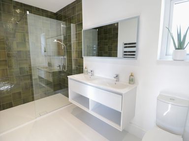 A bathroom with a glass shower enclosure green tiled wall double sinks a mirror and a plant on the windowsill at Bryn Mor in Benllech