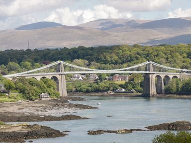A suspension bridge over a river with trees and houses in the background and mountains in the distance