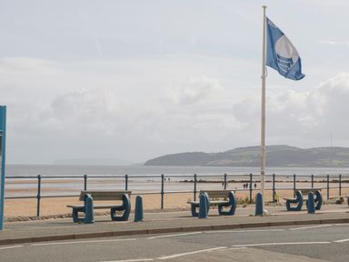 Benches and a flagpole with a flag on a promenade along a sandy beach with hills in the background at Bryn Mor in Benllech