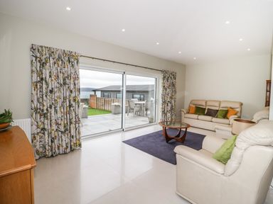 A living room with beige sofas a glass coffee table and floral curtains at Bryn Mor in Benllech