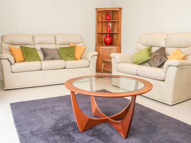 A living room with two beige sofas with cushions a wooden glass top coffee table a wooden corner shelf with red vases and a purple carpet at Bryn Mor in Benllech