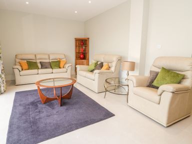 A living room with beige sofas green and orange cushions a glass coffee table a side table with a lamp a wooden corner cabinet and a purple rug at Bryn Mor in Benllech