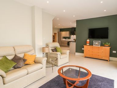 A living room with beige sofas and colorful cushions and a glass coffee table and a wooden sideboard with a TV mounted on a green wall at Bryn Mor in Benllech