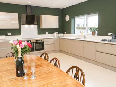A kitchen with beige cabinets green walls a wooden dining table with flowers and a bottle at Bryn Mor in Benllech
