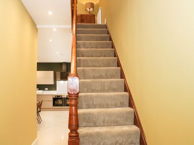 A carpeted staircase with wooden banister leading to a hallway with a lamp and cupboard and a kitchen area visible to the left at Bryn Mor in Benllech
