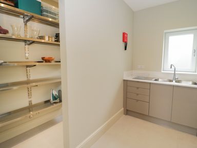 A utility room with a double sink and cabinets next to metal shelving with kitchen items at Bryn Mor in Benllech