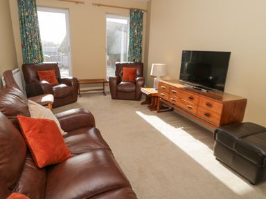 A living room with brown leather sofa and armchairs a wooden TV stand with a flat screen TV and a table lamp at Bryn Mor in Benllech