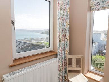 A corner with two windows showing a sea view a wooden chair a radiator and patterned curtains at Bryn Mor in Benllech