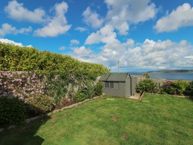 A garden with a small shed surrounded by plants and a hedge overlooking a body of water at Bryn Mor in Benllech