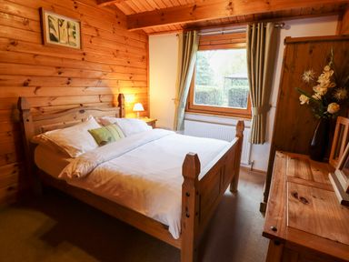 A bedroom with a wooden bed a window with curtains a bedside table with a lamp a wooden dresser with a vase of flowers and a framed picture on the wall at Avocet Lodge in Tattershall Lakes Country Park