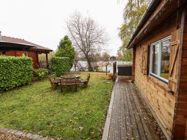 A garden with wooden benches and table between two wooden lodges near a lake at Avocet Lodge in Tattershall Lakes Country Park
