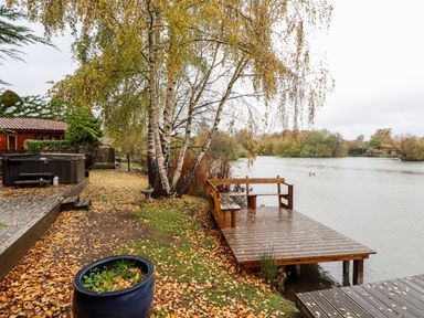A wooden dock with bench by a lake and a cabin with hot tub nearby at Avocet Lodge in Tattershall Lakes Country Park