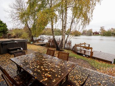 An outdoor wooden table with chairs on a deck and a wooden dock by a lake with trees at Avocet Lodge in Tattershall Lakes Country Park