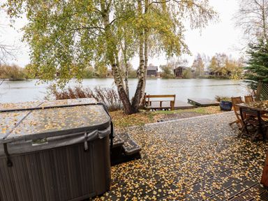 A wooden deck with a hot tub and outdoor furniture next to a lake with trees at Avocet Lodge in Tattershall Lakes Country Park