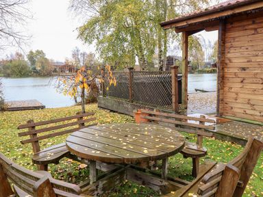 A wooden picnic table with benches on grass near a lake next to a wooden cabin at Avocet Lodge in Tattershall Lakes Country Park