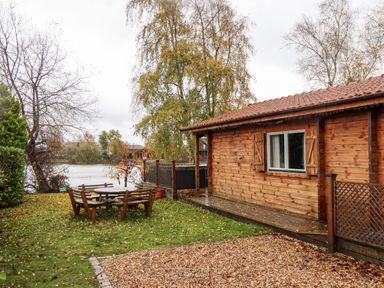 A wooden cabin next to a lake with outdoor bench and table set at Avocet Lodge in Tattershall Lakes Country Park