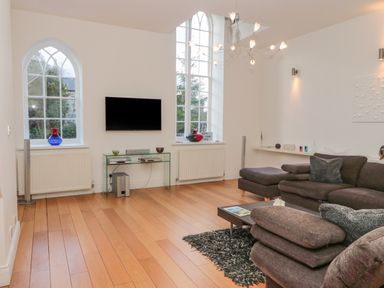 A living room with two arched windows a wall mounted TV and brown sofas at 2 The Reformed Church in Embleton