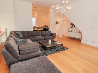A living room with a sectional sofa and coffee table on a rug and a kitchen area in the background at 2 The Reformed Church in Embleton