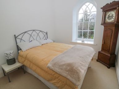 A bedroom with a bed a side table lamp an arched window and a grandfather clock at 2 The Reformed Church in Embleton