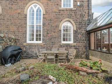An outdoor patio area with a wooden table and chairs set against a stone wall with arched windows and a covered barbecue at 2 The Reformed Church Embleton