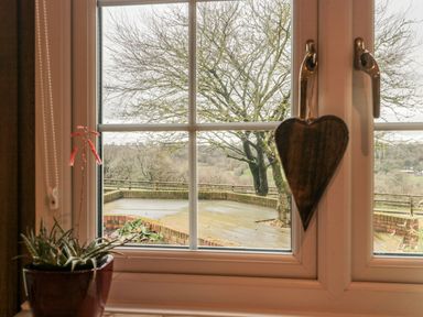 A window with a potted plant and a wooden heart decoration overlooking a patio and trees at Rainbow View in Churchstanton near Churchinford