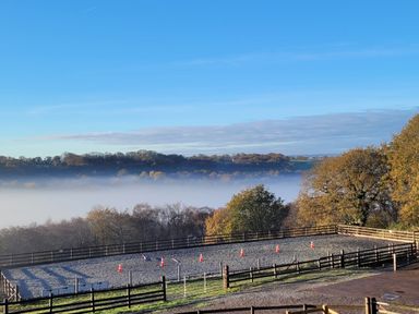 An outdoor horse riding arena with traffic cones surrounded by wooden fences and trees with fog and hills in the background at Rainbow View in Churchstanton near Churchinford